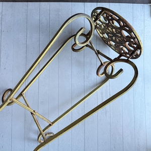 May include: An antique-style plant stand with a circular, ornate top and curved, decorative legs. The metal stand has a distressed, aged appearance with a gold and brown colour scheme. The stand is designed to hold a potted plant.