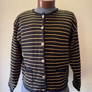 Peut inclure: Un cardigan à rayures noires et dorées avec des boutons dorés et deux poches avant. Le pull est présenté sur un mannequin. Le pull a un col rond et des manches longues.