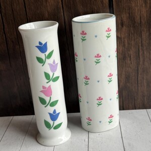 May include: Two white ceramic vases. One vase features a pattern of blue, pink, and purple tulips with green leaves. The other vase has a pattern of pink flowers and blue hearts. Both vases have a glossy finish.