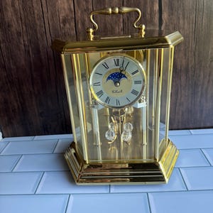 May include: A gold-toned, rectangular clock with a handle, glass panels, and a round clock face. The clock face features Roman numerals and a moon phase indicator. Decorative crystal pendulums hang inside. The clock sits on a white tiled surface.