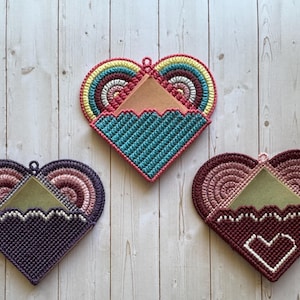 May include: Three heart-shaped, handmade pot holders in shades of purple, teal, and burgundy. Each has a unique design, including a rainbow, a zigzag pattern, and a white heart. They are made of yarn and have a loop for hanging.
