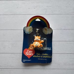 May include: An attachable Friend Bear keychain. The orange bear figure is attached to a blue card with a rainbow and clouds design. The card features the Care Bears logo and text that reads "Fun...festive... attaches to anything!"