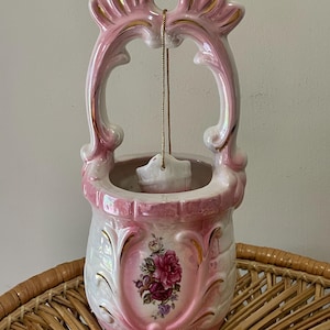 May include: A pink and white ceramic wall hanging in the shape of a well with a gold trim and floral design. The well has a rope and bucket hanging from the top.