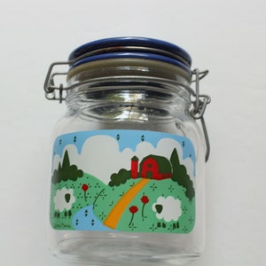 May include: A clear glass jar with a blue lid and a white, green, red, and yellow farm scene decal. The decal features a red barn, green trees, a yellow path, and white sheep.
