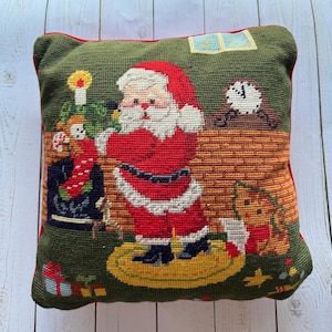 Puede incluir: Un cojín cuadrado con temática navideña con fondo verde y borde rojo. El cojín presenta un diseño de punto de aguja de Papá Noel, un reloj, un árbol de Navidad y un perro. El cojín está sobre una superficie de madera clara.