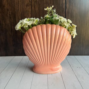 Może przedstawiać: Brzoskwiniowy wazon w kształcie muszli wypełniony białymi i zielonymi kwiatami. Wazon ma żebrowany wzór i stoi na białej drewnianej powierzchni. Tło to ciemnobrązowa drewniana ściana.