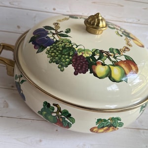 Puede incluir: Cazuela de esmalte crema con tapa, asas y pomo dorados. El plato está decorado con un diseño colorido de frutas y vides alrededor del borde. Las asas y el pomo son de color dorado.