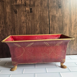 May include: A rectangular, red decorative planter with gold accents. The planter features gold leaf-like designs and rests on four gold claw feet. The interior is red, and the rim is trimmed with gold. The background is a dark wood.