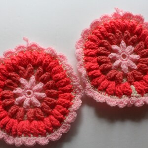 Puede incluir: Dos posavasos de ganchillo en tonos rosa y rojo. Los posavasos son redondos y presentan un diseño de flor en el centro.