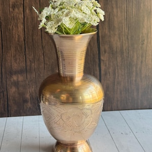 May include: A brass vase with a wide, flared top and a bulbous body. The vase features an etched floral design around the middle and is filled with white flowers. The vase is approximately 12 inches tall.