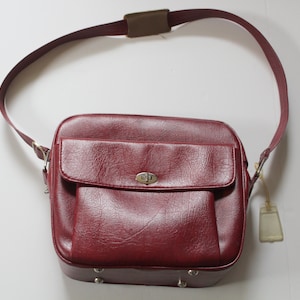 Pode incluir: Uma bolsa de ombro de couro bordô com fecho de aba e fivela prateada. A bolsa tem uma alça única e um pequeno bolso na frente.