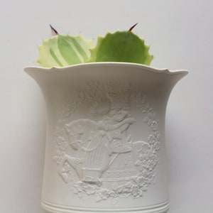 Puede incluir: Maceta de cerámica blanca con un diseño en relieve de una mujer montando un caballo de carrusel. La maceta tiene un borde festoneado y está llena de una planta suculenta verde.