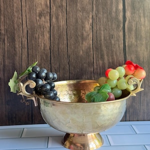 May include: A gold-colored, hammered metal fruit bowl with a pedestal base. The bowl is filled with artificial grapes in shades of green, red, and black. The bowl has decorative handles on each side. The background is a wooden wall.