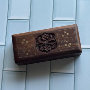 May include: A rectangular wooden box with a dark brown finish. The box features intricate floral carvings and gold-colored accents. The design includes a central carved floral motif and decorative corner patterns.