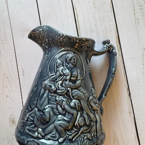 Puede incluir: Una jarra de cerámica azul con un diseño en relieve de una mujer y niños. La jarra tiene una base ancha y un asa con un adorno decorativo.