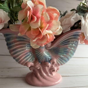 Może przedstawiać: Różowy ceramiczny wazon w kształcie motyla z tęczowymi skrzydłami. Wazon jest wypełniony różowymi i białymi kwiatami.