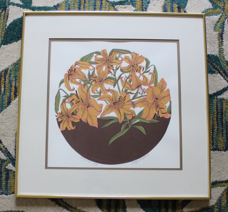 May include: A framed print of a circular design featuring a cluster of orange lilies with brown and green accents. The print is titled "Lily and Lilies" and signed by the artist.