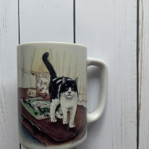 Puede incluir: Taza de cerámica blanca con una ilustración de un gato blanco y negro. El gato está sentado en una mesa con una lámpara y un teléfono. La taza tiene un asa en el lado derecho.