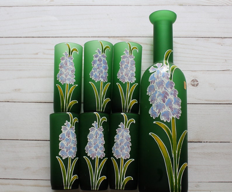 Puede incluir: Una botella de vidrio verde y seis vasos de chupito a juego. Cada pieza est&aacute; decorada con un dise&ntilde;o floral que presenta una flor de lavanda con hojas verdes y detalles amarillos. El conjunto se exhibe sobre una superficie de madera clara.