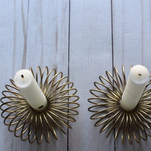 Op de afbeelding: Twee gouden metalen kaarsenhouders met een spiraalvormig ontwerp. Elke houder heeft een witte kaars erin.