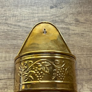 May include: A vintage brass wall-mounted container with a hinged lid. The container features an embossed grape vine design around the sides. The top has a curved shape with a hole for hanging. The container is made of shiny brass.