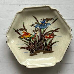 May include: Cream-colored, hexagonal decorative plate with a gold rim. The plate features a floral design with blue, orange, and red irises, along with brown and green foliage. The plate has a crackle glaze finish.