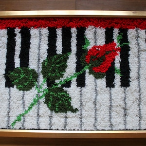 以下が含まれることがあります： 赤、白、黒のピアノの鍵盤のデザインが施された額縁付きの敷物。緑色の茎に葉と赤いバラが鍵盤の上に置かれています。