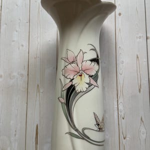 May include: A tall, off-white ceramic vase with a flared, taupe-colored rim. The vase features a pink and cream orchid design with gray and black accents, and a butterfly. A decorative piece for the home.