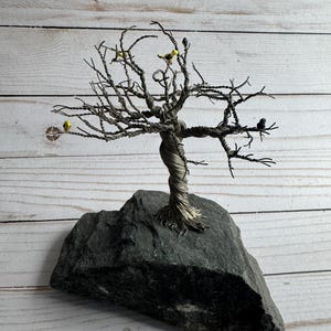 May include: A handcrafted wire tree sculpture with a twisted trunk and delicate branches, mounted on a dark gray stone base. The tree features small, yellow and black bird figurines perched on its branches, creating a nature-inspired art piece.