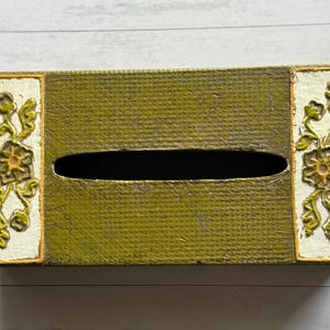 May include: Rectangular tissue box with a textured olive green surface. The box features floral relief designs in yellow and white on each end. The opening for tissues is an oval shape.