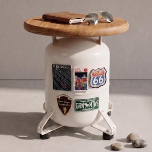 May include: A side table constructed from a white cylindrical metal tank base with four angled legs and a round wooden top. The tank is decorated with various travel-themed stickers including Route 66 and Green Mountains. A leather notebook and sunglasses rest on top.