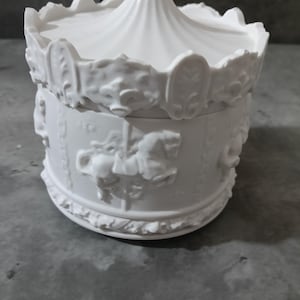 Può includere: Scatola portagioie bianca in ceramica a tema giostra. La scatola presenta un design dettagliato di cavalli della giostra ed elementi decorativi. Il coperchio è a forma di tendone da circo. È un oggetto decorativo.