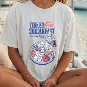 May include: A cream-colored t-shirt with the words "TURKISH BREAKFAST" in blue and red, featuring a graphic of a breakfast platter. The shirt is worn with a navy blue cap with white anchor designs.