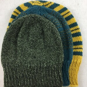 Puede incluir: Tres gorros de punto. El primero es verde oscuro con motas blancas, el segundo es verde azulado y el tercero es verde azulado con rayas amarillas. Los gorros están hechos de un hilo suave y texturizado.