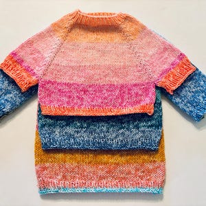 May include: Three hand-knitted sweaters in different sizes are stacked. The top sweater is pink and orange, the middle one is pink and blue, and the bottom one is blue, yellow, and orange. They have long sleeves and a round neckline.