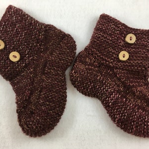 Puede incluir: Un par de botitas de bebé de punto marrón con cierres de botón de madera.