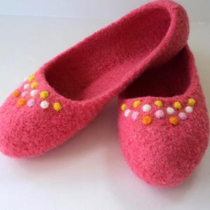 May include: A pair of bright pink felted wool slippers featuring a rounded toe design. The front of each slipper is decorated with a pattern of small, raised dots in yellow, white, and orange colors, creating a textured and colorful appearance.