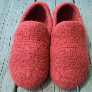 Puede incluir: Un par de zapatillas sin cordones confeccionadas con un material de fieltro grueso y texturizado en un tono rojo anaranjado vibrante. Las zapatillas presentan un diseño de punta redondeada, una abertura profunda y una estructura sólida.