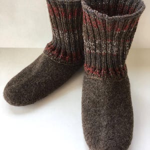 May include: A pair of dark brown felted wool slippers featuring a high, ribbed knit cuff. The cuffs are detailed with vertical stripes in dark brown, white, and red yarn. The slippers have a rounded toe and a solid, textured felted wool body.