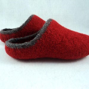 May include: A pair of red felted wool slippers featuring a rounded toe design and a dark grey trim along the opening. The slippers have a textured surface and a slip-on style, with one slipper positioned slightly behind the other in the frame.