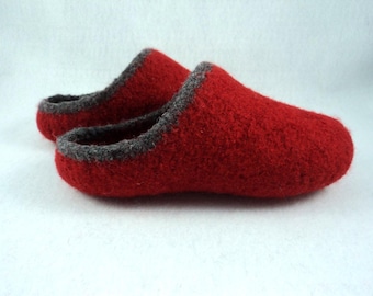 May include: A pair of red felted wool slippers featuring a rounded toe design and a dark grey trim along the opening. The slippers have a textured surface and a slip-on style, with one slipper positioned slightly behind the other in the frame.
