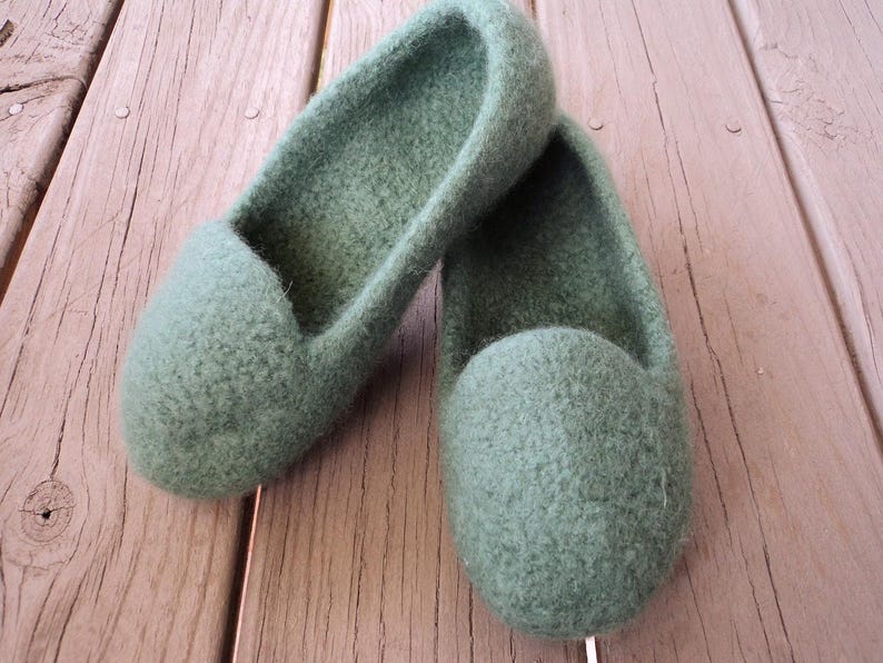 Puede incluir: Un par de zapatillas de lana afieltrada en color verde salvia con punta redondeada y dise&ntilde;o sin cordones. Las zapatillas presentan un material grueso y texturizado con una superficie suave y difusa y una abertura profunda para el pie.