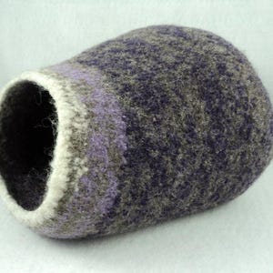 Pode incluir: Um recipiente em forma de taça feito de lã feltrada, com uma superfície texturizada e mesclada em tons de roxo profundo, cinzento e castanho. A abertura circular é rematada com uma banda grossa e contrastante de lã cor de creme.