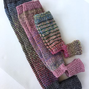 Peut inclure: Trois paires de jambières tricotées à la main avec des bords-côtes en haut et en bas. La première paire est gris anthracite foncé, la deuxième est un mélange mauve et rose, et la troisième est un mélange multicolore de bleu, vert et jaune.