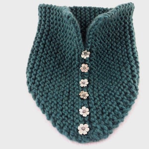 May include: A dark teal knitted neck warmer featuring a vertical row of six silver-toned flower-shaped buttons down the centre. The accessory is crafted with a chunky knit texture and has a slightly pointed bottom edge, creating a distinct shape.