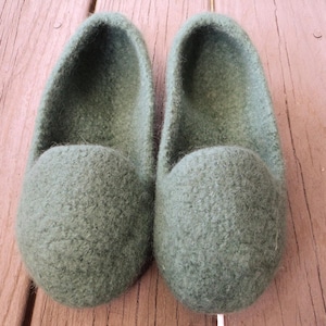 Puede incluir: Un par de zapatillas de lana afieltrada de color verde oliva con dise&ntilde;o de punta redondeada y estilo sin cordones. Las zapatillas presentan una superficie densa y texturizada y una abertura profunda, con un color s&oacute;lido uniforme.