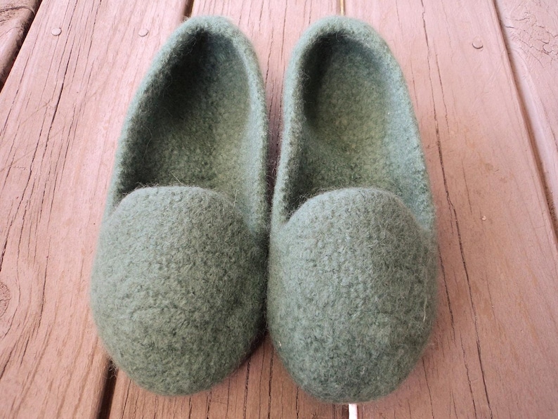 Puede incluir: Un par de zapatillas de lana afieltrada de color verde oliva con dise&ntilde;o de punta redondeada y estilo sin cordones. Las zapatillas presentan una superficie densa y texturizada y una abertura profunda, con un color s&oacute;lido uniforme.