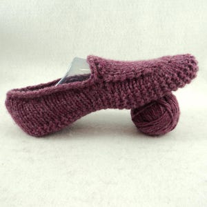 Op de afbeelding: Een pruimkleurige gebreide slipper met een gestructureerd breipatroon, voorzien van een ronde neus en een lage opening. Een bijpassende bol pruimkleurig garen is onder het hielgedeelte van de slipper geplaatst voor ondersteuning.