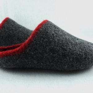 May include: A pair of dark grey felted wool slippers featuring a rounded toe design and a vibrant red trim along the collar and opening. The slippers have a textured, dense surface and a slip-on style with a low-profile silhouette and no visible heel.