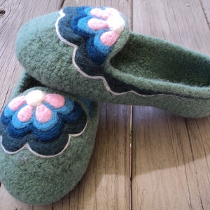 Puede incluir: Un par de zapatillas de lana afieltrada en color verde salvia con una aplicaci&oacute;n floral festoneada en la parte superior. El dise&ntilde;o de la flor consta de p&eacute;talos superpuestos en tonos rosa claro, azul brillante y azul marino, con un borde blanco.